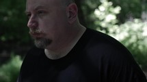 Sad, anxious, depressed looking Christian man in black shirt with beard in nature walking, hiking, meditating, praying contemplating in green, wooded area in trees with sunlight shining in cinematic, slow motion.