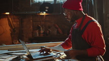 African American Blacksmith Drawing Sketch and Using Laptop in Workshop