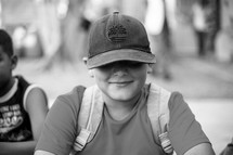 Black and white portrait of a school boy with a book bag and ball cap smiling.