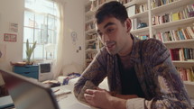 Young handsome man smiling, chatting on video call on laptop when sitting at desk in sunlit room, working or communicating online at home. Medium shot
