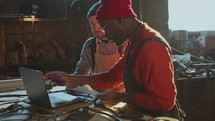 Blacksmith Discussing Metalwork Project on Laptop with Colleague in Workshop