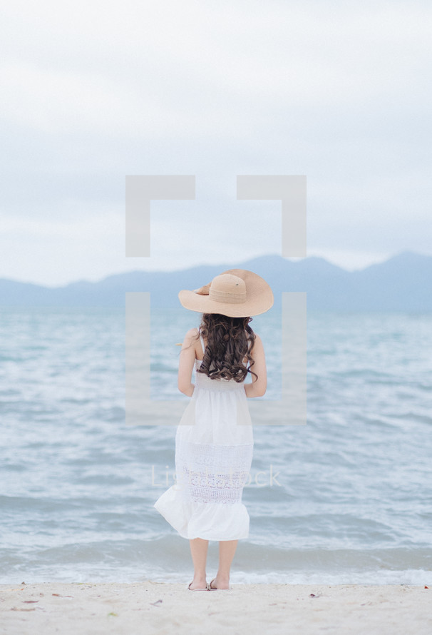 a woman in a white dress and sunhat on a shore 