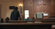 Young man in black suit walking into old church building with stained glass window sits down on pew.