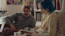 Young man lying on bed with guitar, discussing book with his girlfriend with blue hair, spending the day together at home. Gen Z couple, relationships concept
