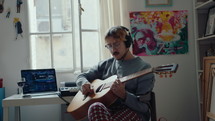 Young musician in headphones sitting by desk with audio equipment, playing the guitar, singing a song, recording music at home studio during the day
