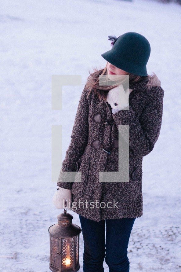 woman in a winter coat walking in snow carrying a lantern 