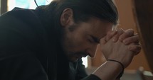 Young, emotional, anxious, and stressed man with long hair and black suit sitting in old church in worship and praying.