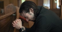 Young, emotional, anxious, and stressed man with long hair and black suit sitting in old church in worship and praying.