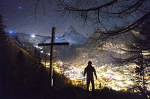 man standing next to a cross looking down at lights in a valley 