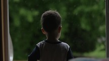 A young, cute boy, child stands inside watching the rain fall outside in cinematic slow motion.