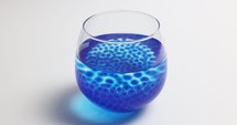 Growing Blue Hydrogel Balls In A Glass Of Water Against White Background. - Closeup Shot