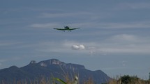Light propeller plane flies in France, featuring tall grass, mountains, and blue sky in the frame. It's a practice flight of the propeller plane, flying towards and exiting the frame