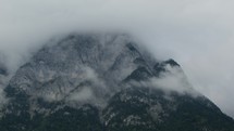 A mountain with grey rocky textures partially hidden by thick white clouds against a cloudy sky, with wooded foothills visible at the foot