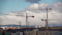 An urban landscape with construction cranes, buildings and structures, mountains in the background, and flying clouds