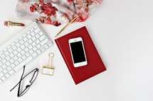 red lipstick, reading glasses, clip, computer keyboard, phone, scarf, pen, and red book on a desk 