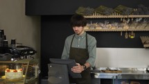 Young cashier in apron typing on touch screen in bakery store. Close up, indoor, slow motion.