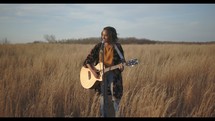 a woman standing in a field holding a guitar singing 