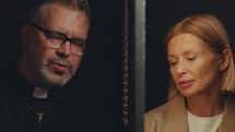 Senior female penitent and priest sitting on both sides of lattice screen and talking in private during confession in Catholic church
