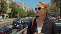 Slow motion - a young woman in a black jacket and white-rimmed sunglasses is walking along the street, holding a mobile phone