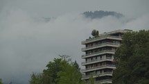 An apartment building with a roof garden stands in the foreground among fog-covered hills and trees