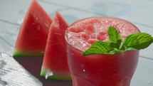 A close-up of watermelon slices and a dessert in a transparent glass on the table, garnished with sprigs of mint, with slow-motion shots of ice cubes falling onto them