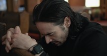 Young, emotional, anxious, and stressed man with long hair and black suit sitting in old church in worship and praying.