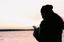 a woman looking at her cellphone standing in front of a calm sea at sunset 