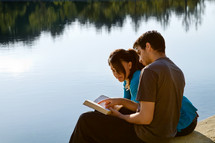 Couple reading the Bible while sitting on a pier at a lake.