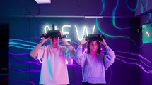 A man and a woman putting on virtual reality goggles in a neon room.