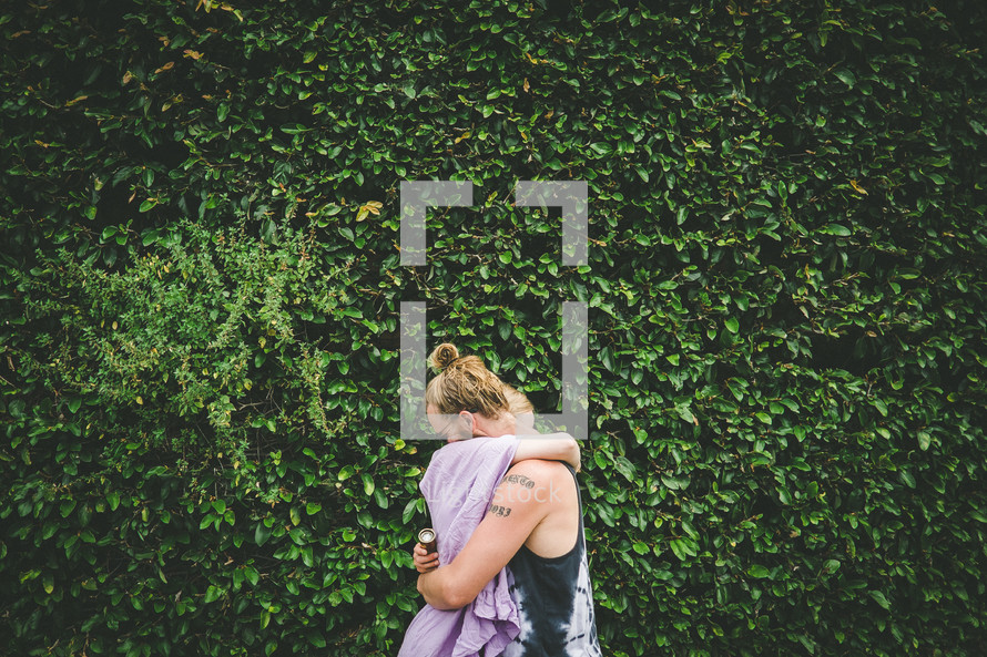 A man and woman embrace in front of a wall of ivy.