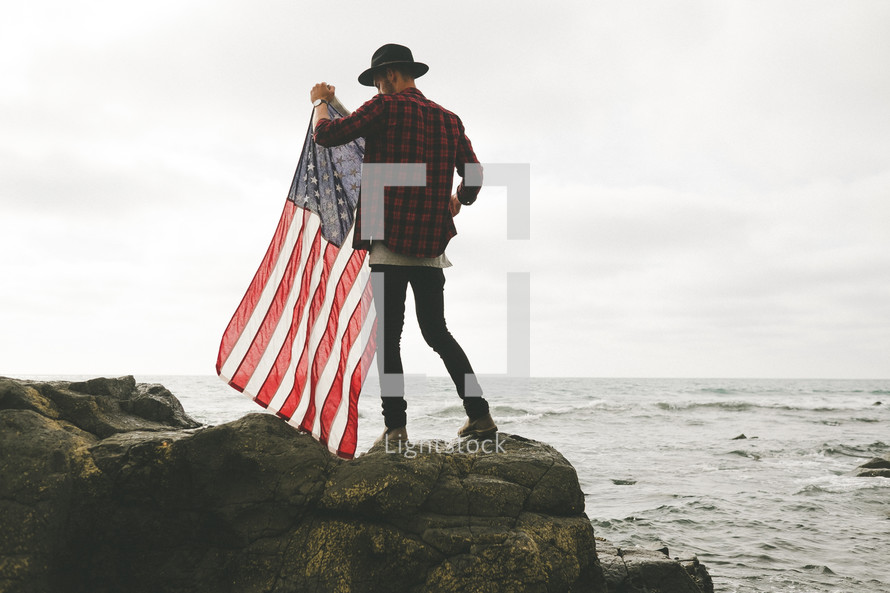 A man stands on a rocky ocean shore with an American flag.
