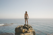 A woman stands on a large boulder overlooking the ocean.