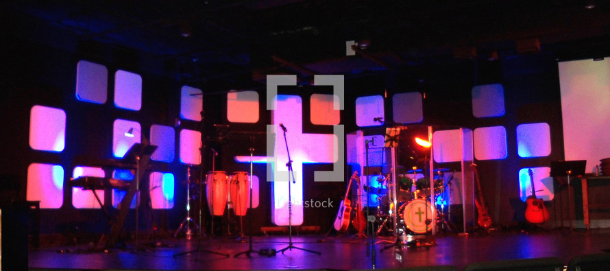 A lighted sound stage for recording music, playing concerts or having a live worship band. The stage is decorated with a cross, lighting, lighted panels and musical instruments including guitars, drums, microphones, bass guitar and other instruments for leading a live praise and worship church service. 