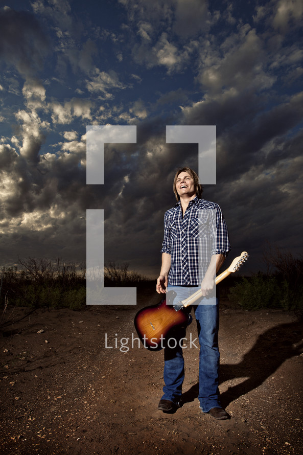 man holding a guitar by the neck under a cloudy sky
