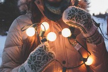 A woman in coat and mittens holds a string of lights.