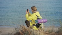 Young man with backpack takes photo of the sea on smartphone while walking along rocky coast.