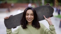 A young brunette girl stands in the background of a park with a skateboard and looks into the camera. Portrait of a girl skateboarder. Spending your leisure time.