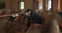 Young, emotional, anxious, and stressed man with long hair and black suit sitting in old church in worship and praying.