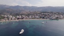 Aerial over turquoise ocean littered with yachts, view of white sand beach and coastal town Himarë in Albania