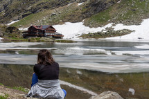 A woman hiking attire sits contemplatively on the lakeshore looking toward a lodge