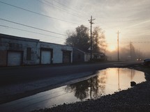 puddle on the side of a rural road and abandoned warehouse buildings at sunrise 