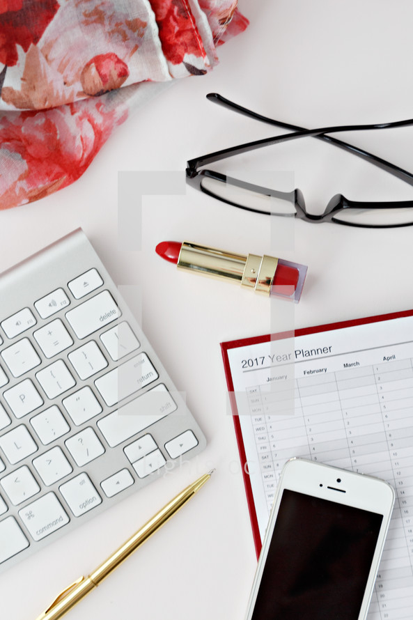 red lipstick, reading glasses, clip, computer keyboard, phone, scarf, pen, and planner on a desk 