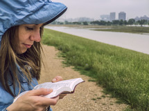 A woman in a rain coat reading the Bible near a river.