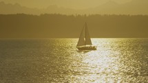 A sailboat passes through golden sunset light on the water in front of Olympic mountain range, slow-motion
