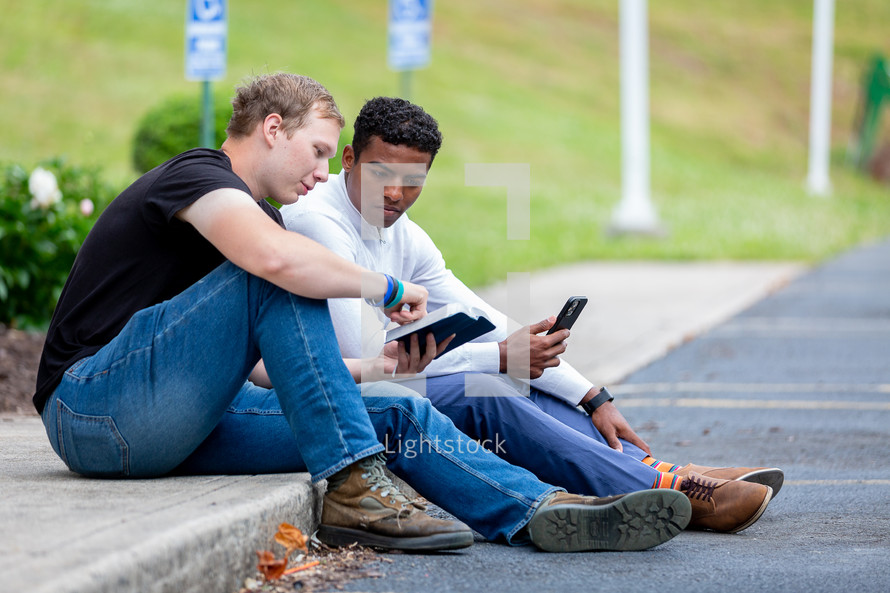 Two men sitting on sidewalk looking at the Bible