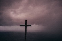 Wooden cross in the ocean water covered by low-lying storm clouds.