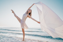 Young beautiful ballerina dancing ballet on seashore with huge silk fabric fluttering in wind.Concept of tenderness, lightness, art and talent in nature.
