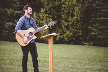 a man playing a guitar outdoors in front of a podium 