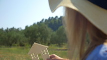 Slow motion of a woman in summer hat holding wooden house model on the background of green forest and blue sky. Concept of eco or country house, real estate and mortgage