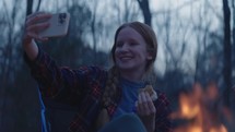 Young man and woman smile and take a selfie beside a camp fire